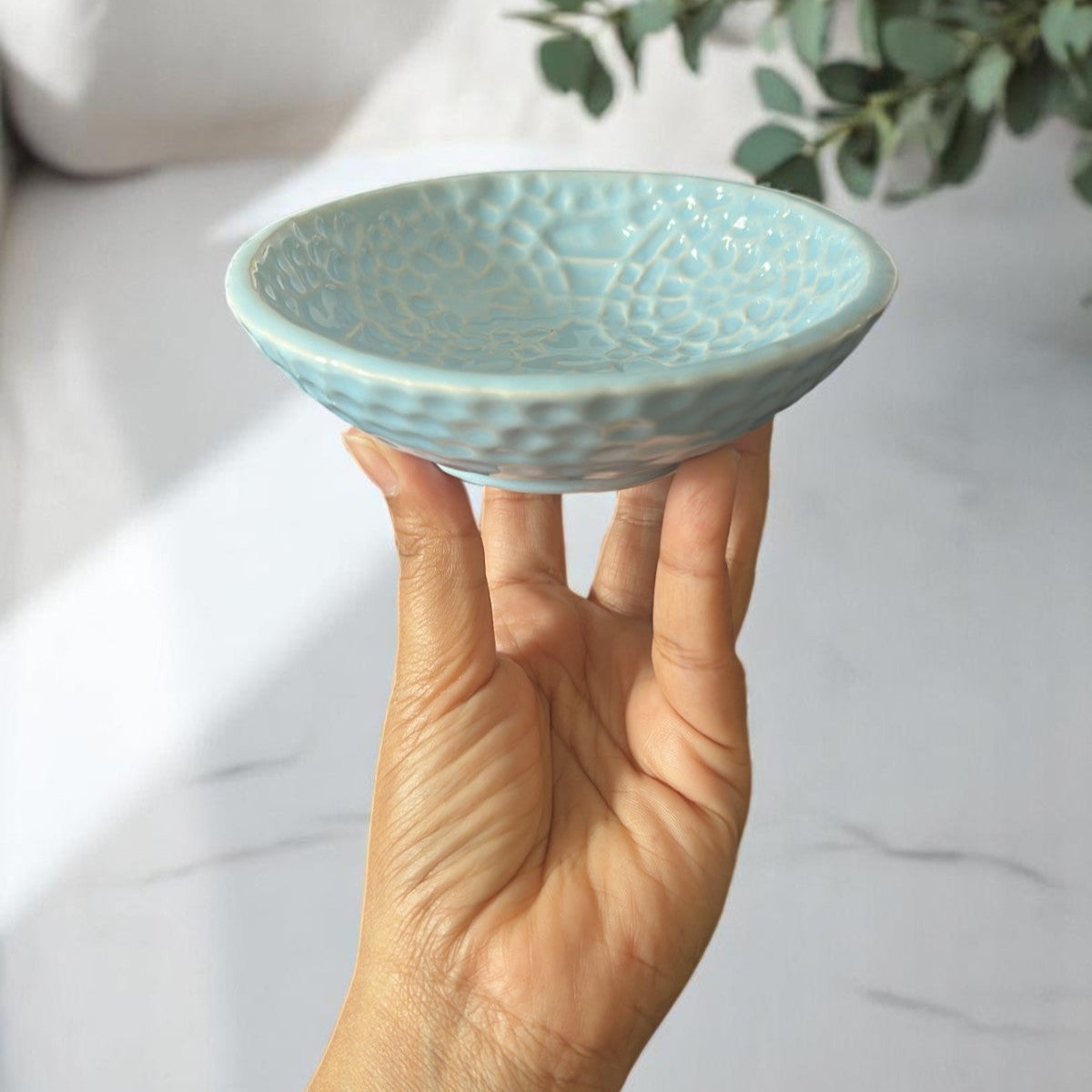Hand holding a small turquoise ceramic bowl with a textured surface against a light background.