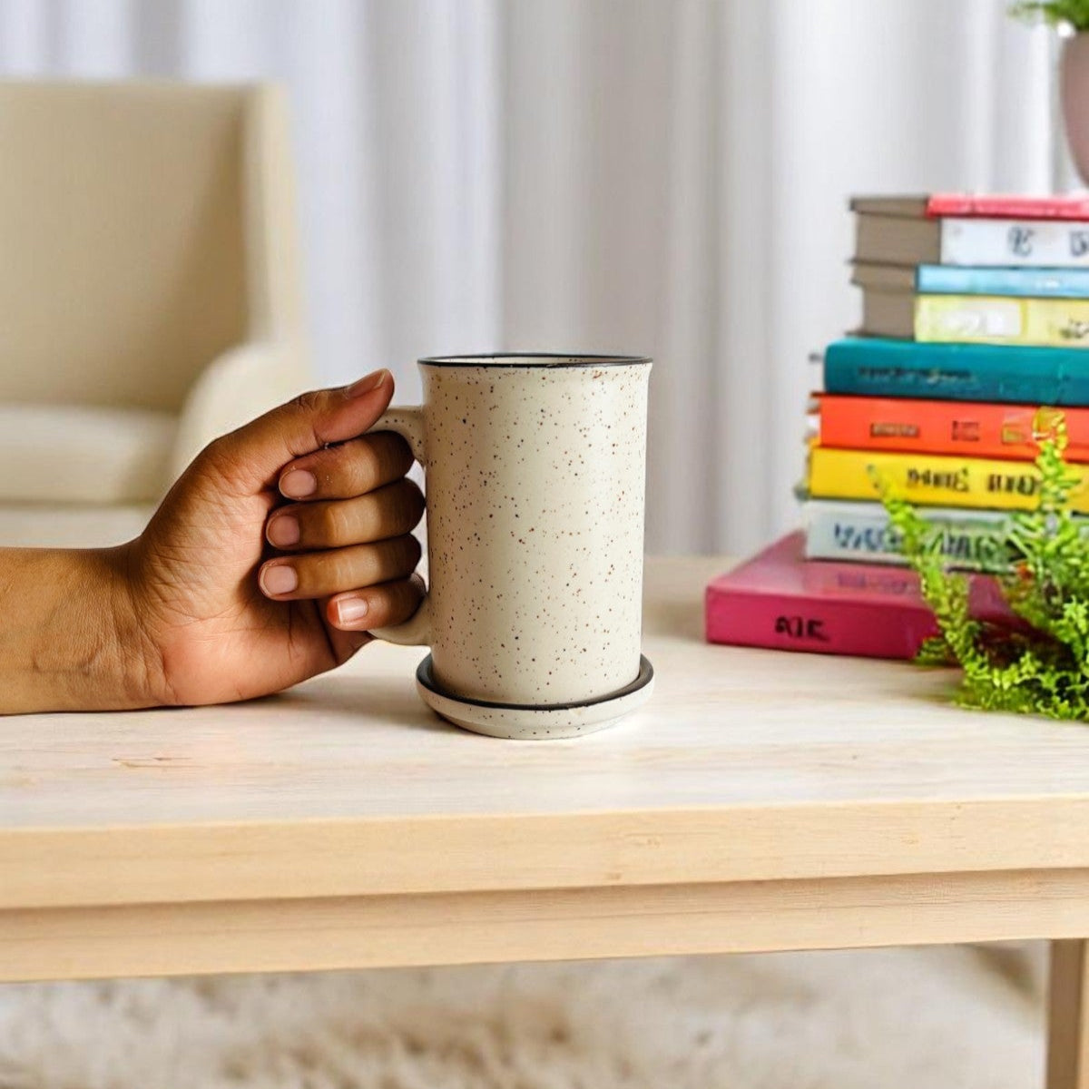 Hand holding a speckled ceramic mug on a wooden surface with books and a plant in the background.