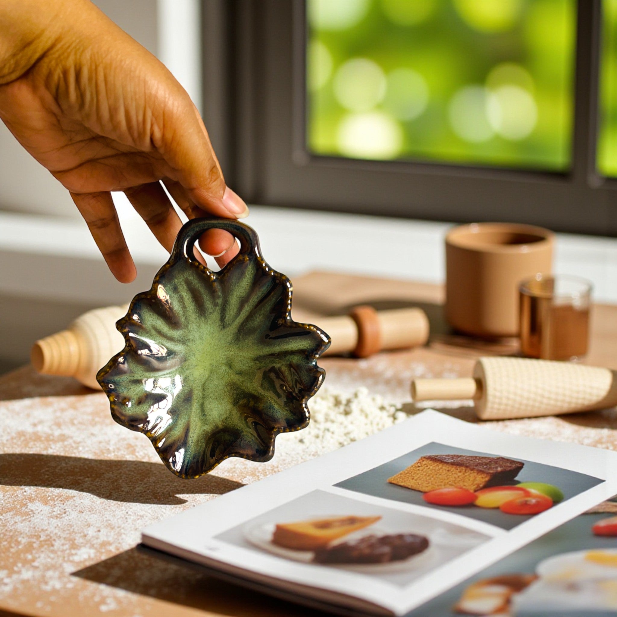 Hand holding a green ceramic leaf tray over a cookbook with baking images on a kitchen counter.