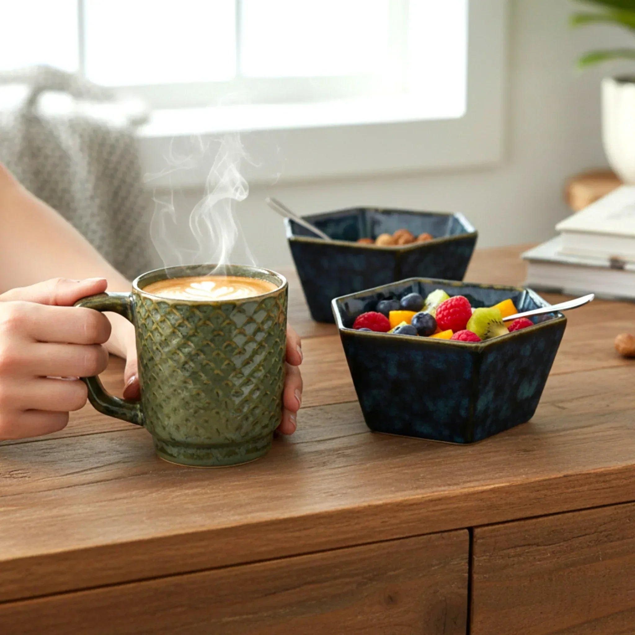 A steaming green mug of coffee on a wooden table with blue ceramic bowls of fruit and cereal laid out for breakfast.