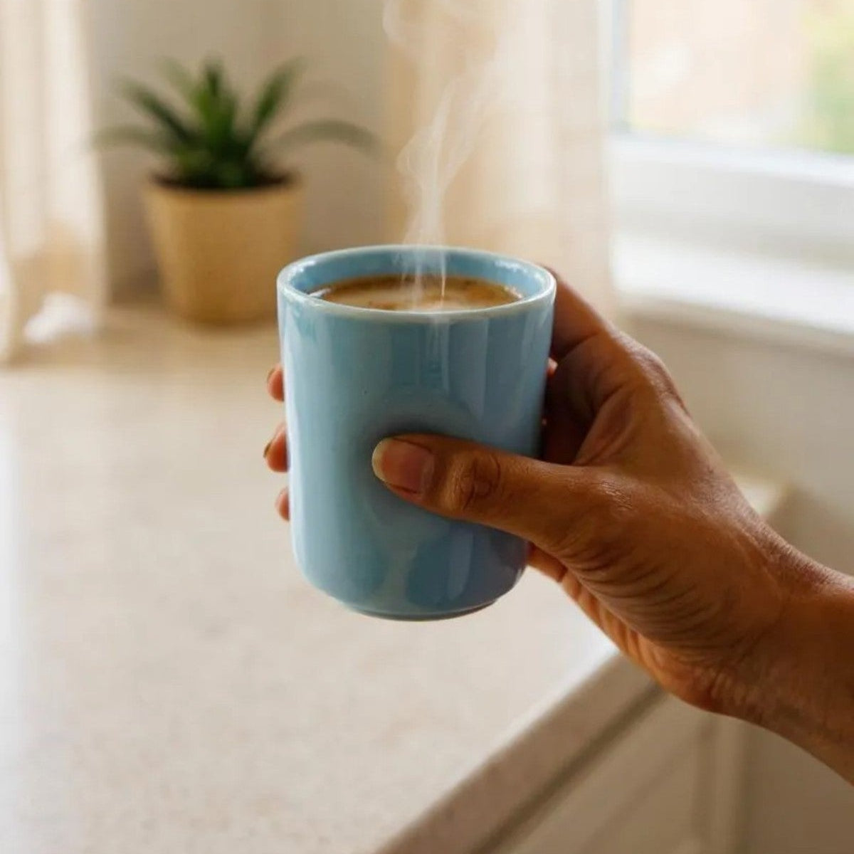 Hand holding a blue mug with steaming coffee near a window