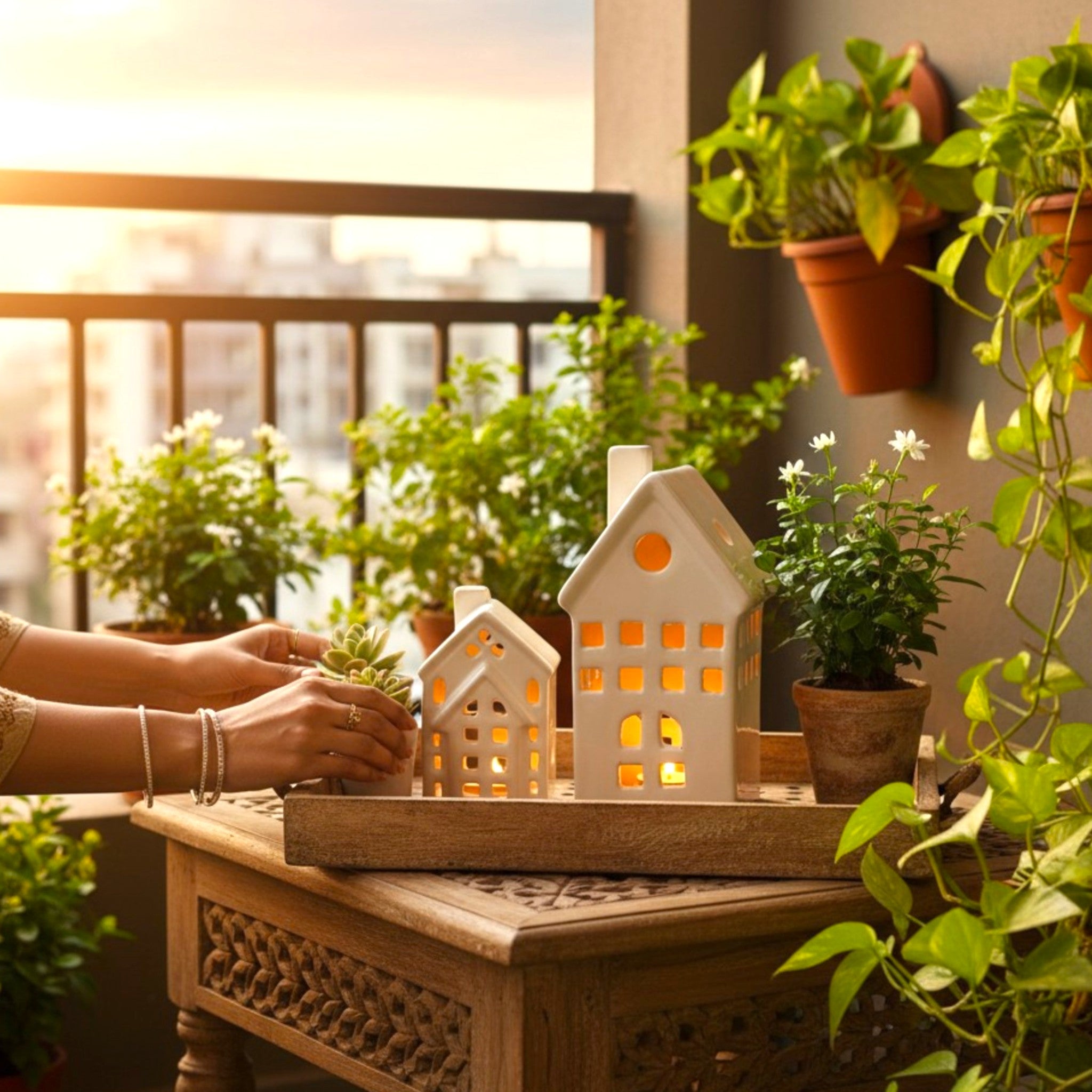 Two white ceramic tea-light houses with warm glowing light inside, sitting on a wooden carved table on a sunlit balcony surrounded by green potted plants.
