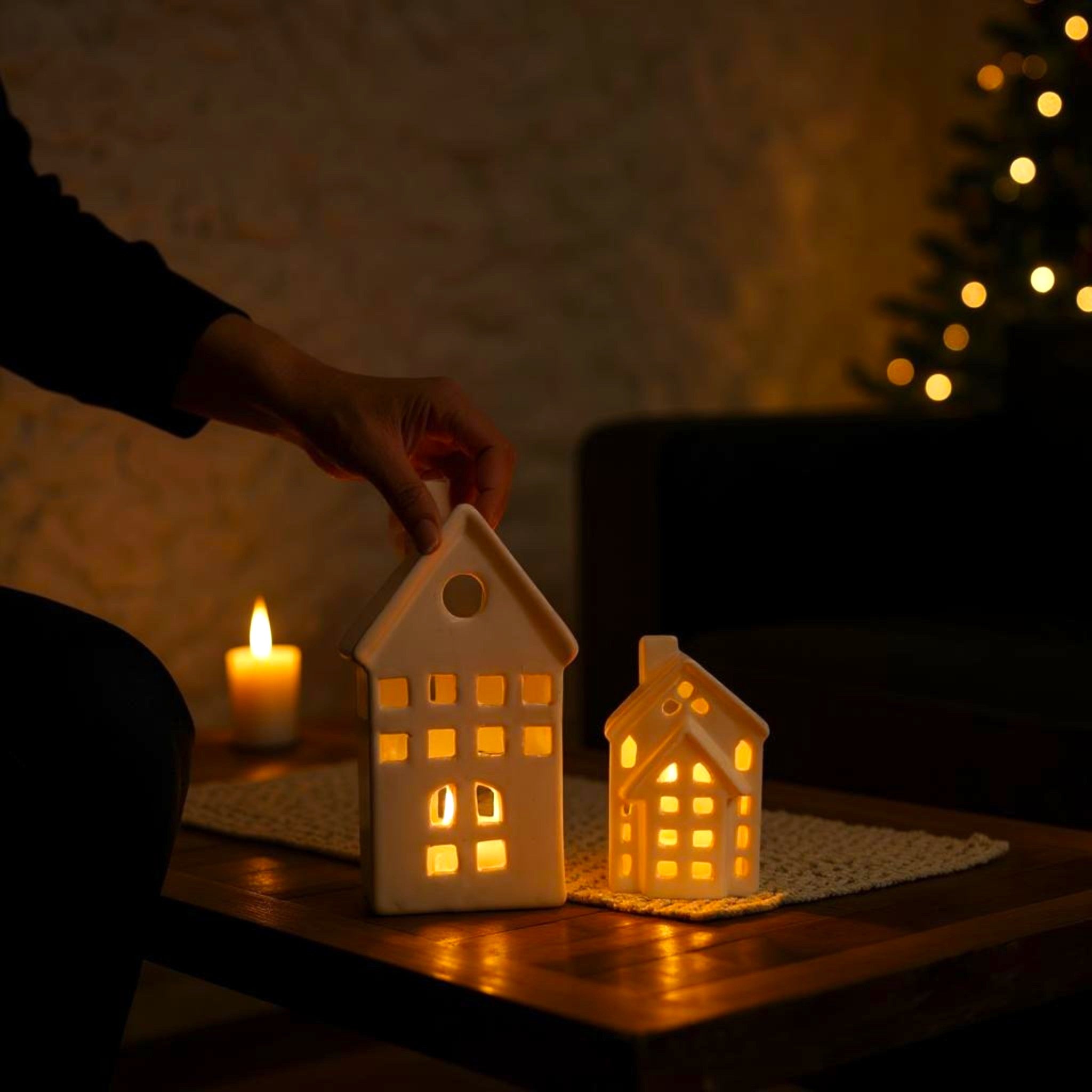 Ceramic house-shaped lanterns being lit on a table with a warm glow, surrounded by soft lighting and a blurred background.