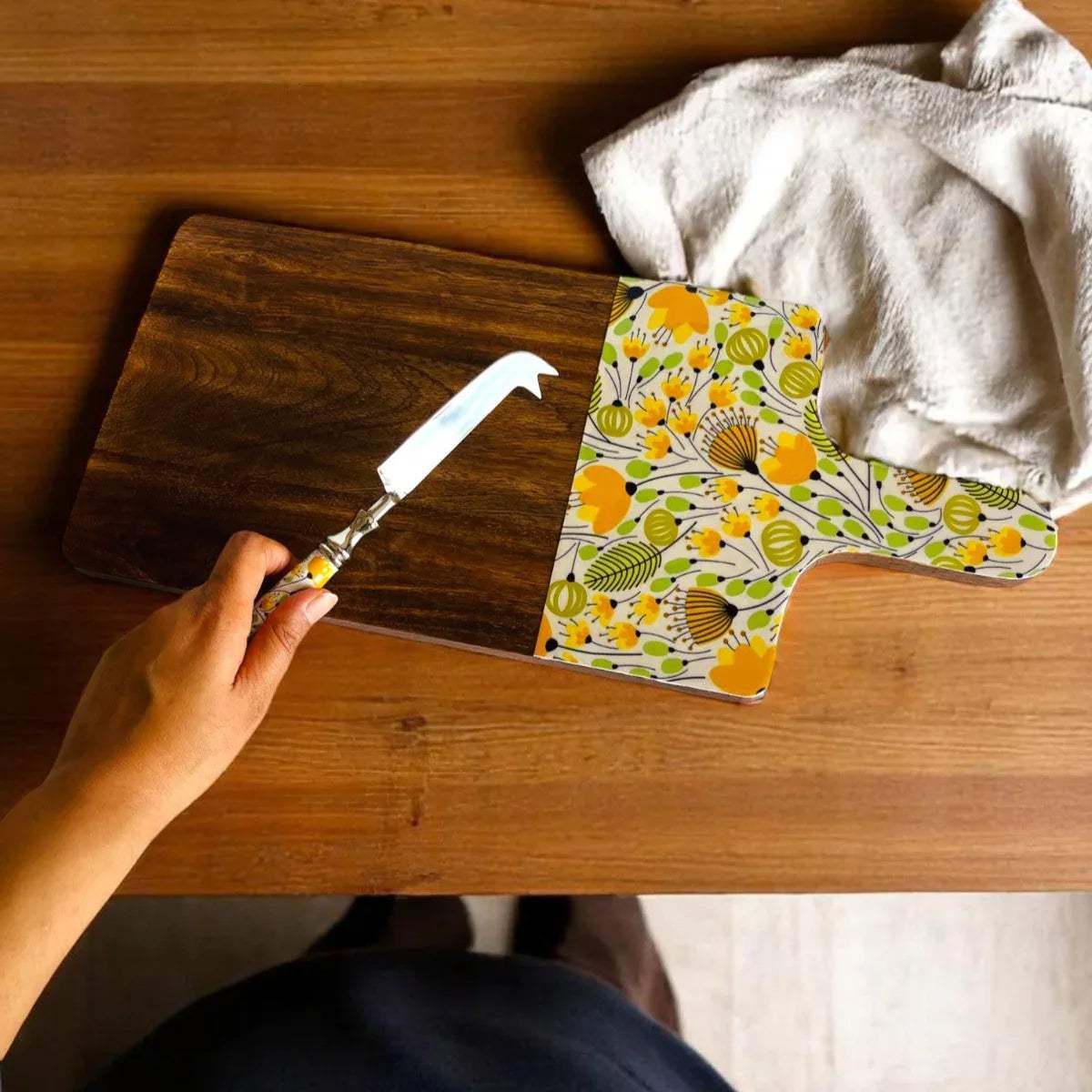 Hand holding a cheese knife over a wooden cheese board with a colorful patterned towel on a wooden surface.