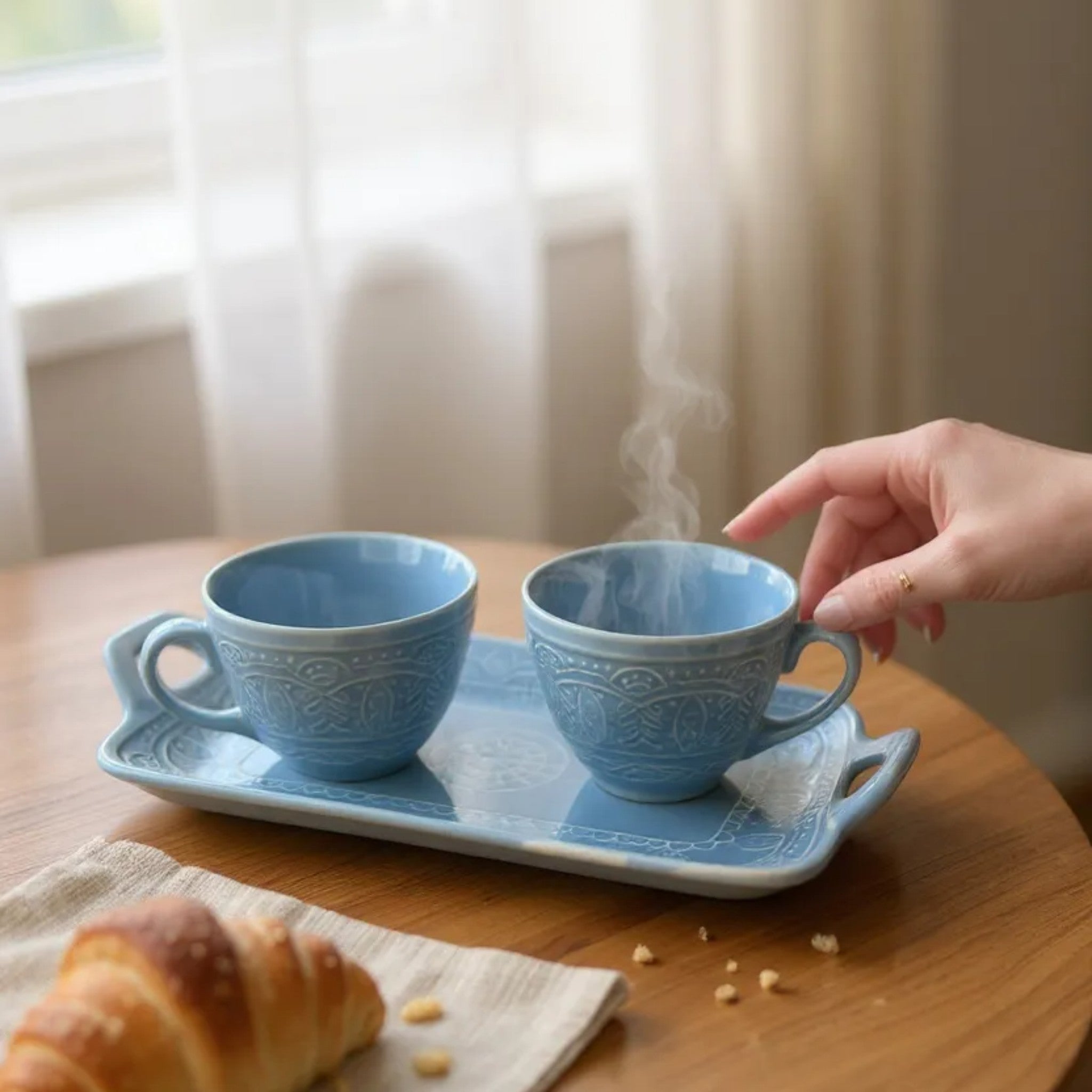 Two blue ceramic cups with steam on a tray, accompanied by a croissant on a wooden table.