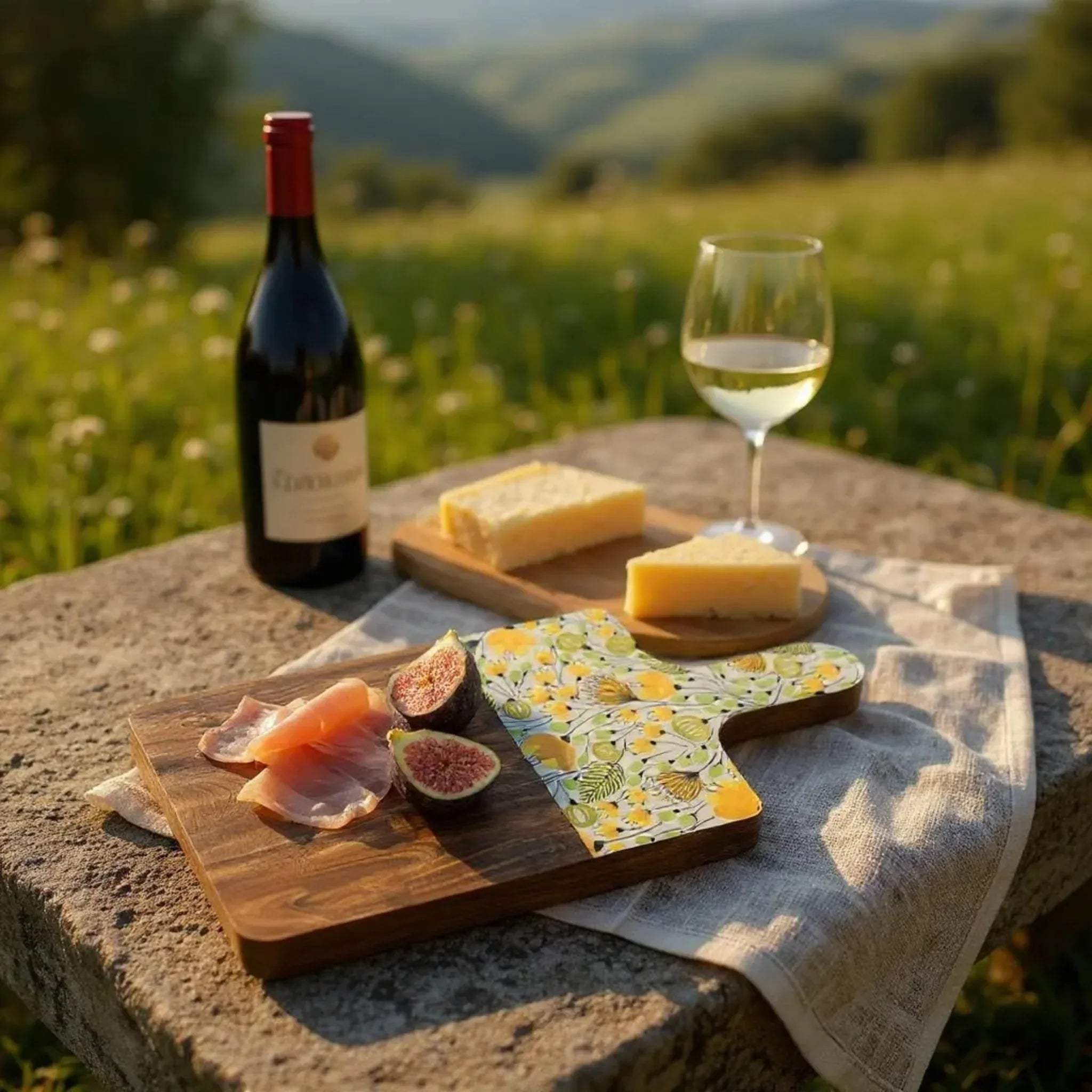 Date setup; the floral board paired with a wine glass and bottle and a loaf of cheese and salmon on a outdoor table.