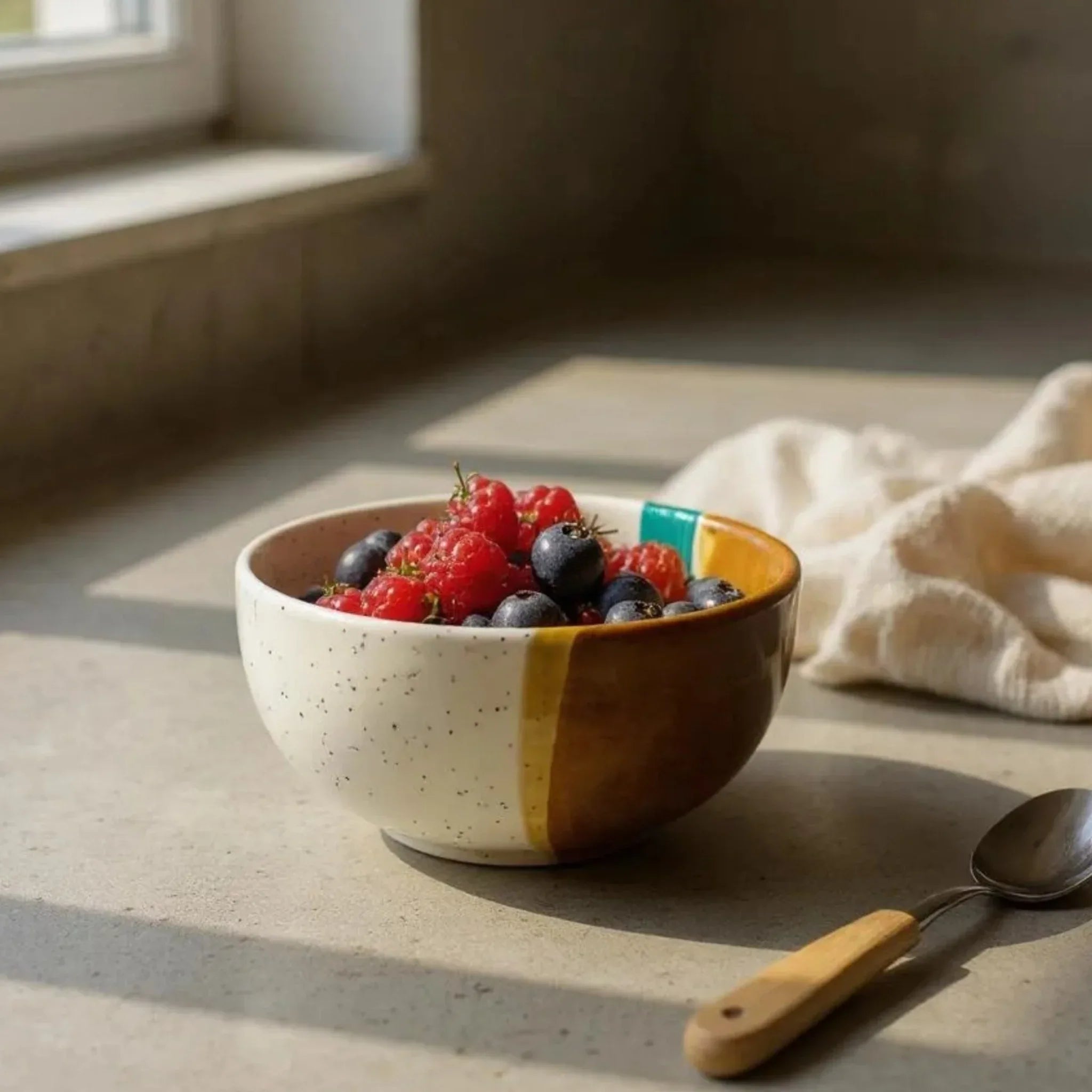 Rustic Stone Kiss of the Coast Bowl with berries on a sunlit countertop, next to a spoon.
