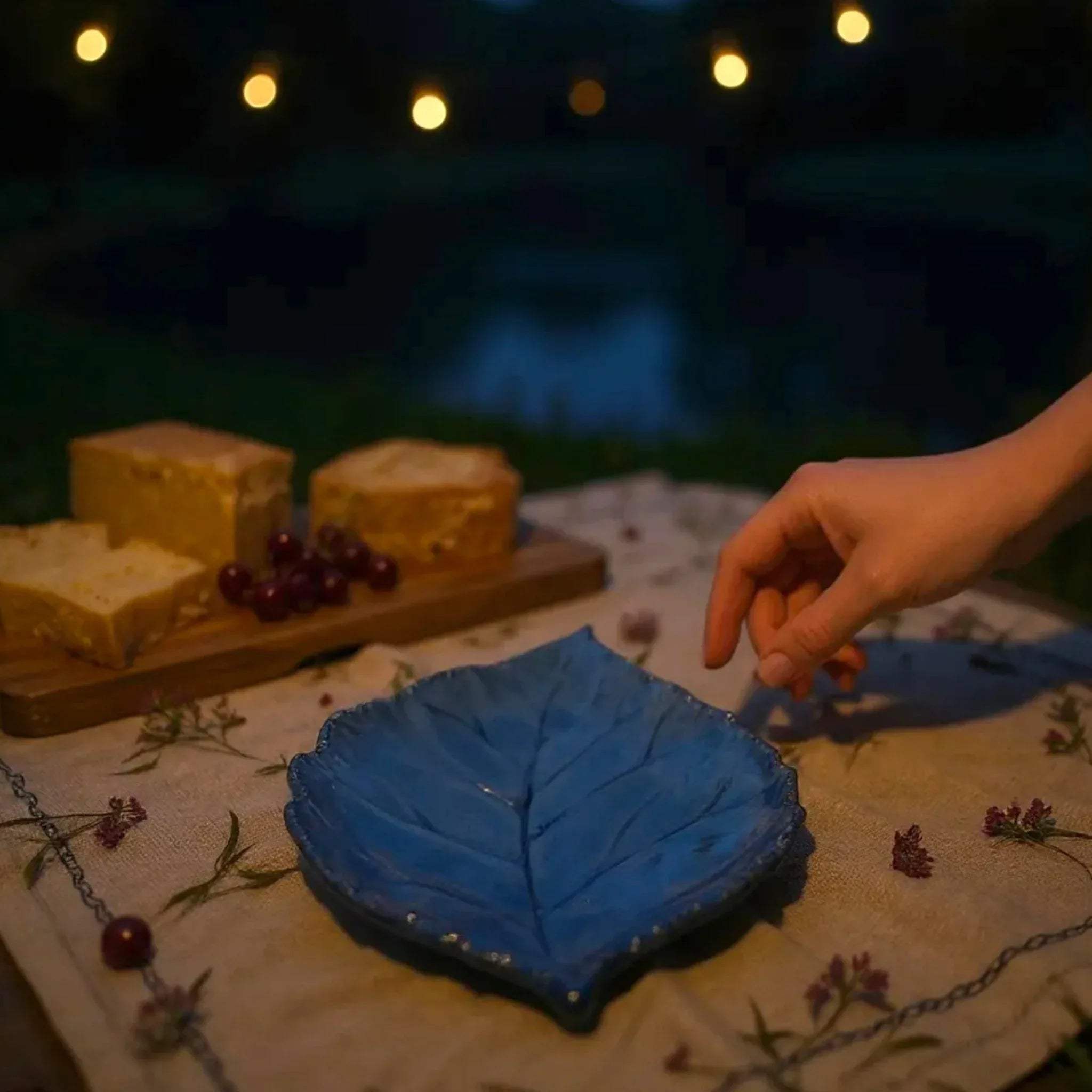 Blue leaf-shaped ceramic platter on a table with string lights in the background in an evening picnic setup.