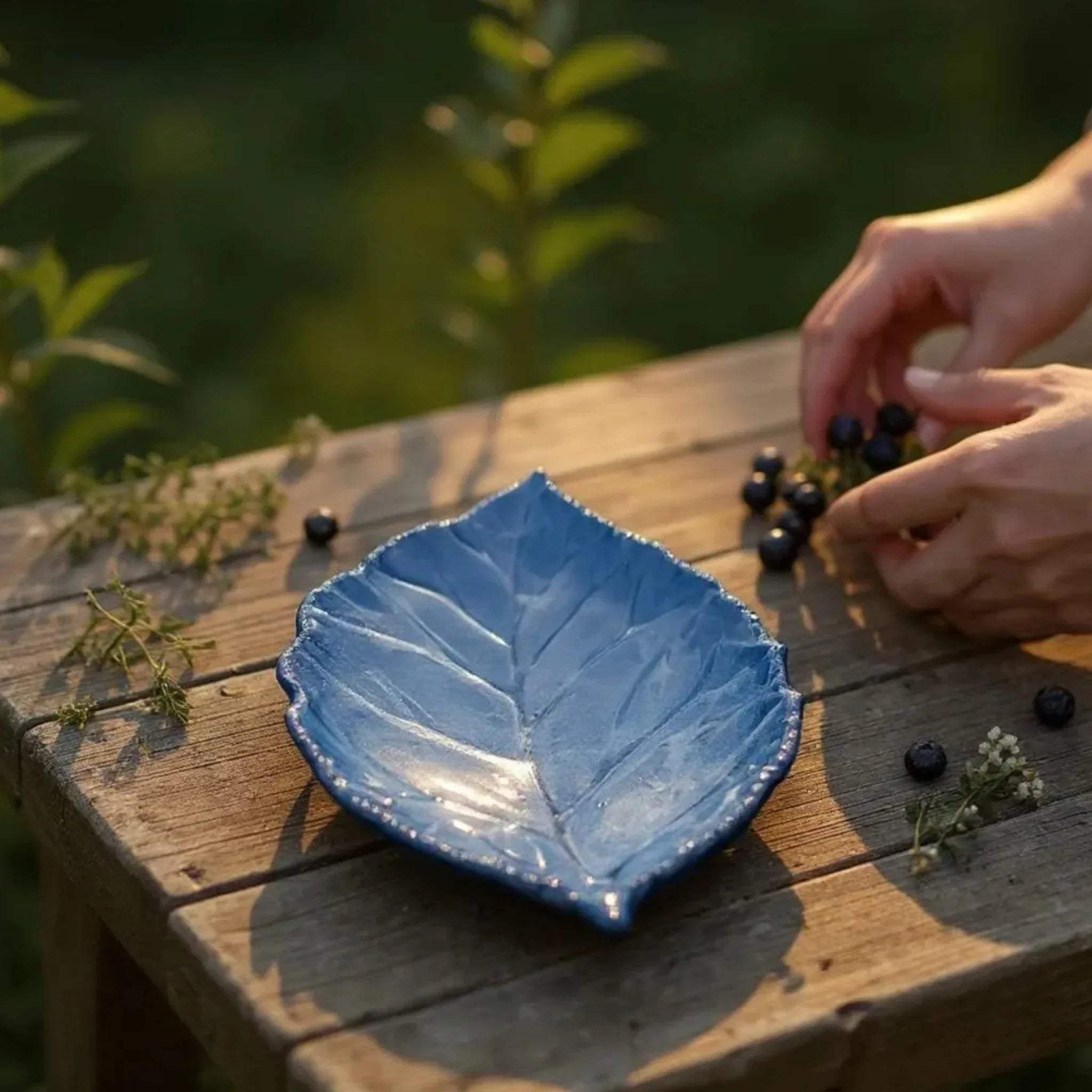 Blue leaf-shaped ceramic plate on a wooden surface with hands arranging berries and wildflower.