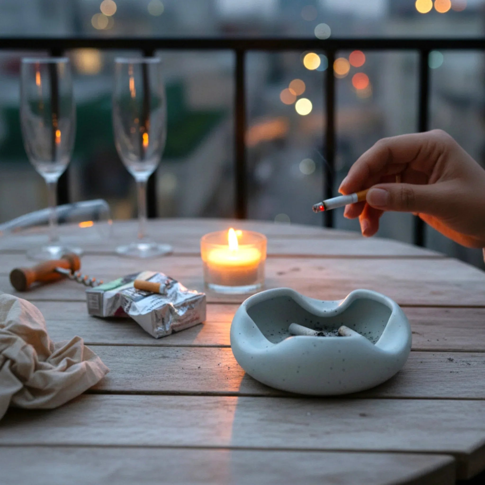 Person lighting a cigarette by a candle on a wooden table with glasses and a grey ashtray.
