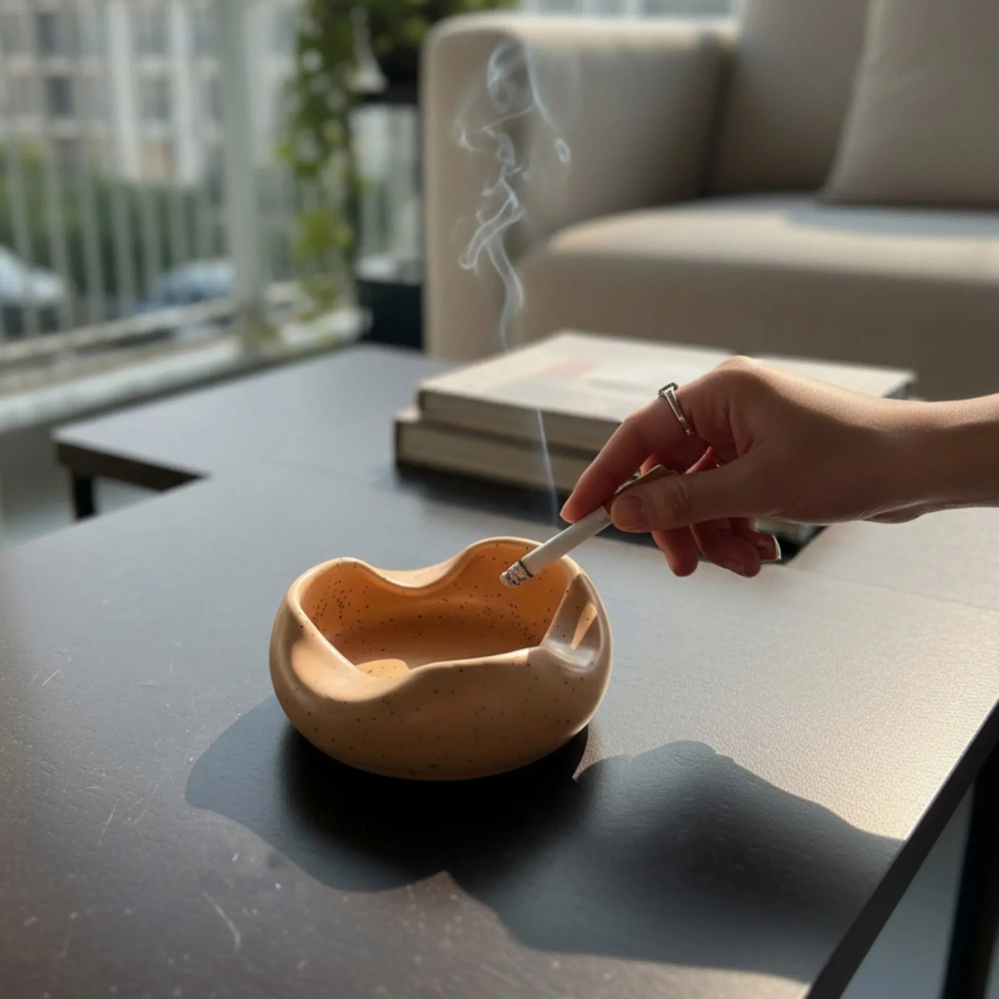 Person lighting a cigarette over an orange / brown / beige cloud-shaped ashtray on a table.