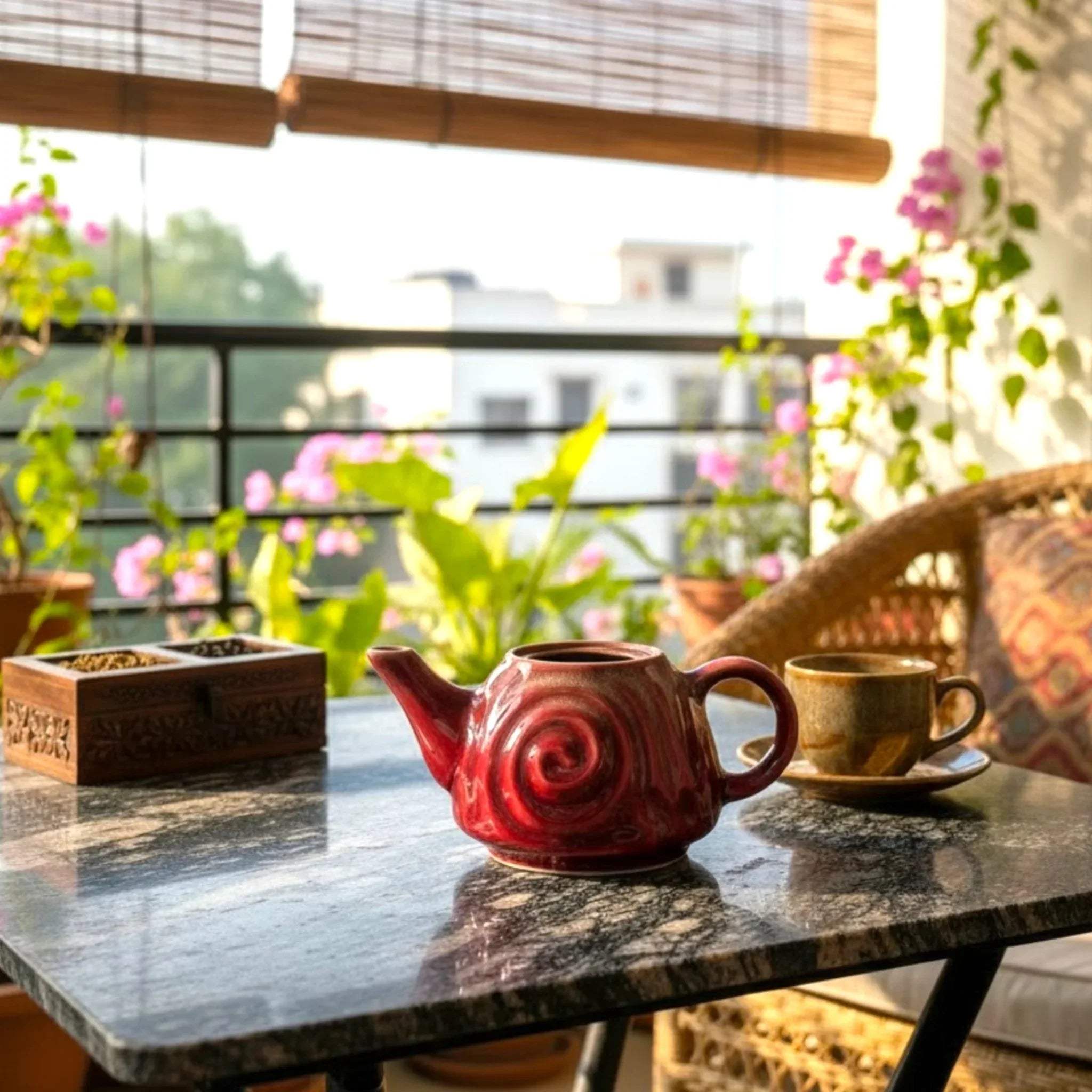 Moody tea set; Ruby Swirl teapot on a marble table in a sunlit balcony with plants around it.