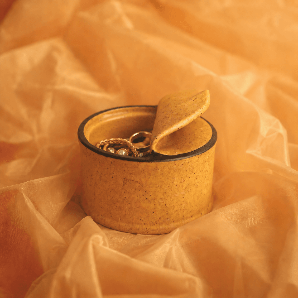 A small round textured ceramic ring box with a hinged lid, shown open and filled with gold rings, set against a background of crumpled warm peach-colored tissue paper.
