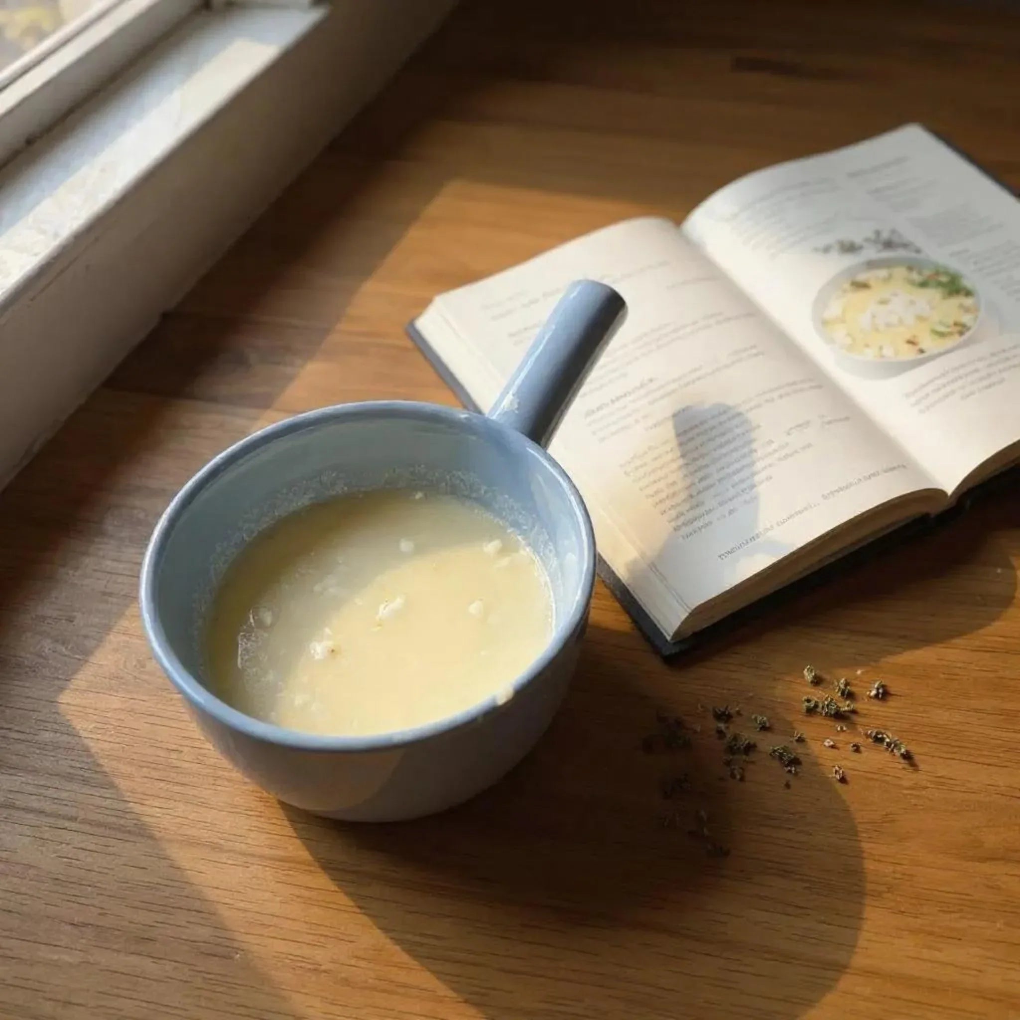 Blue bowl with handle with soup on a wooden table next to an open book.