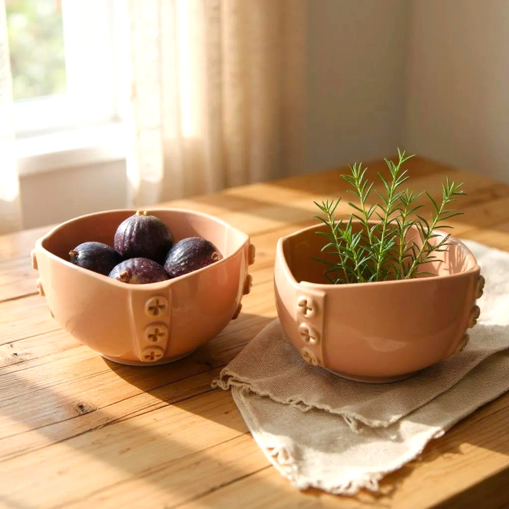 Two pink ceramic bowls on a wooden surface with one containing figs and the other with a plant.