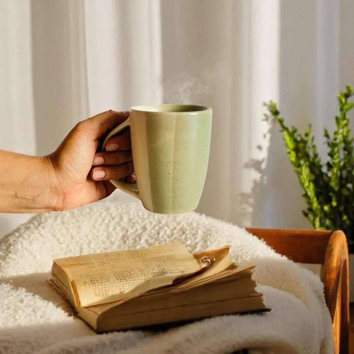 Hand holding a rustic ceramic mug on a cozy blanket with an open book and plant in soft sunlight.