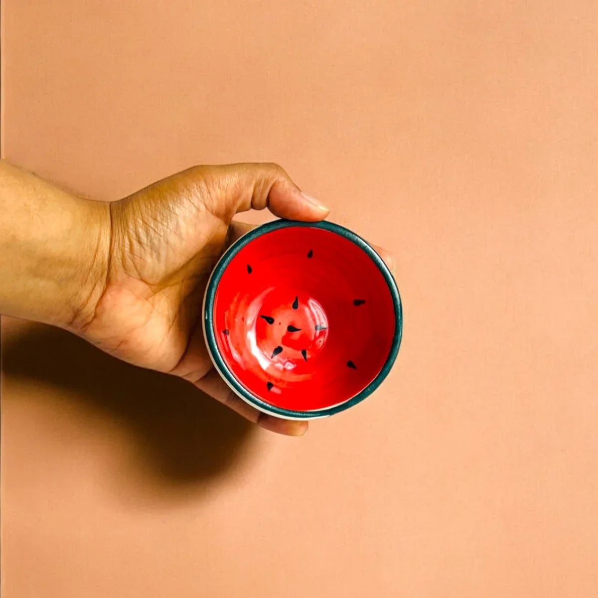Hand holding a red ceramic bowl with black speckles, looking like a watermelon against a beige background.
