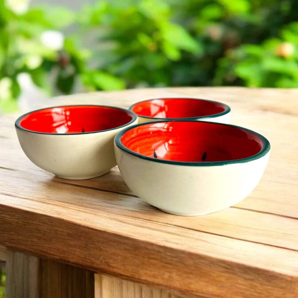 Three red and white ceramic bowls on a wooden surface with greenery in the background