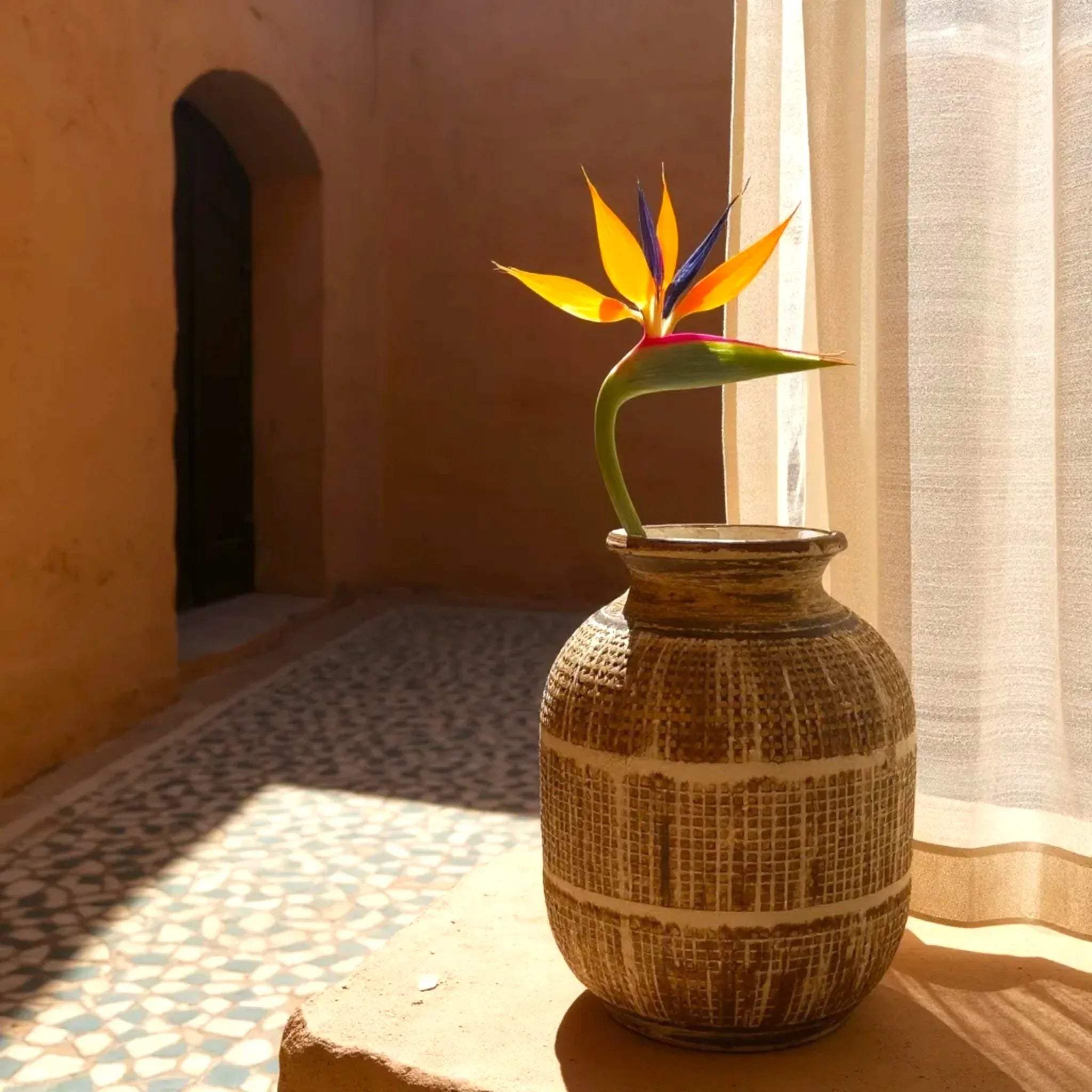 Decorative brown vase with a colorful flower on a stone surface near a window with curtains.